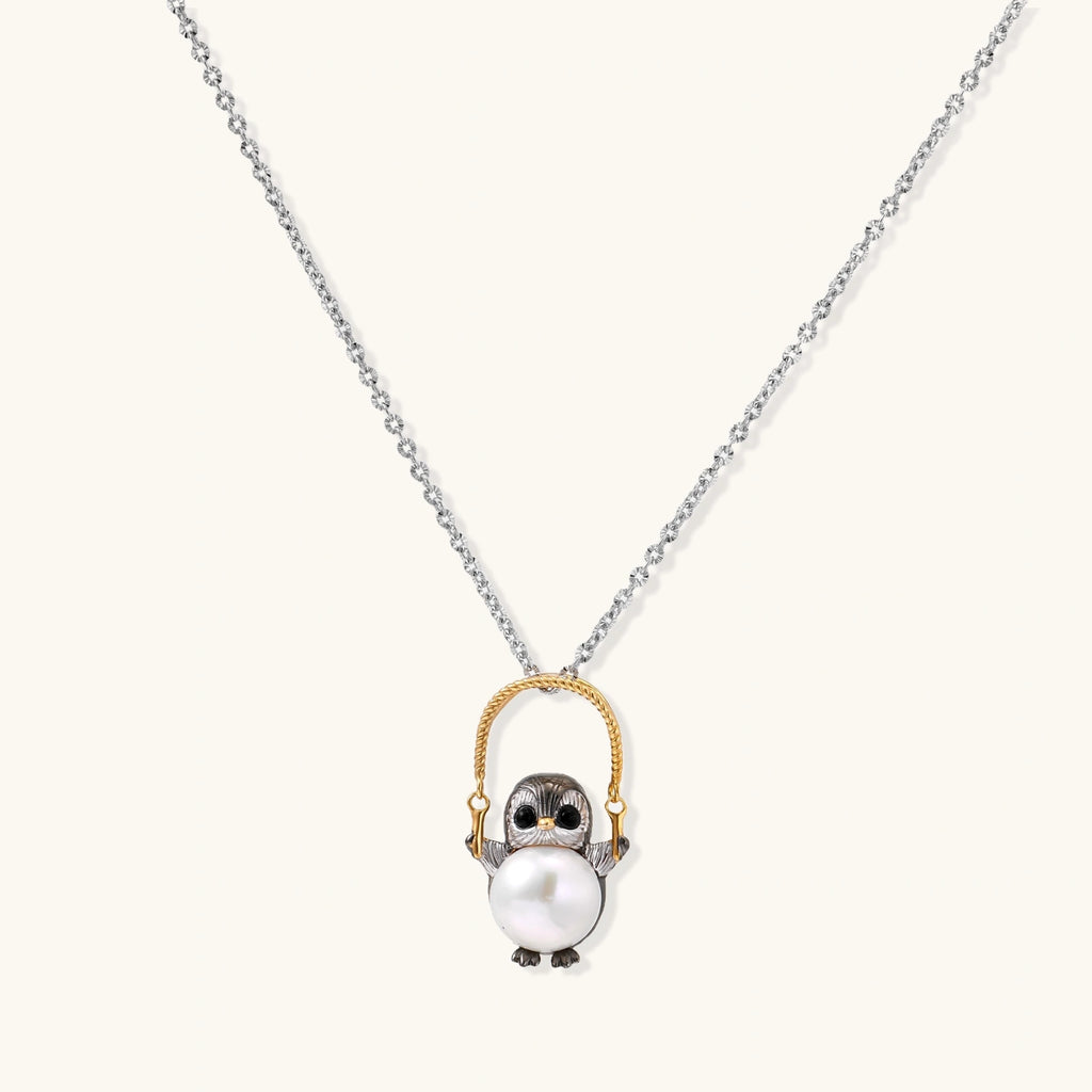 Whimsical penguin pendant holding a white pearl on a gold swing, rhodium-plated silver chain on cream background.