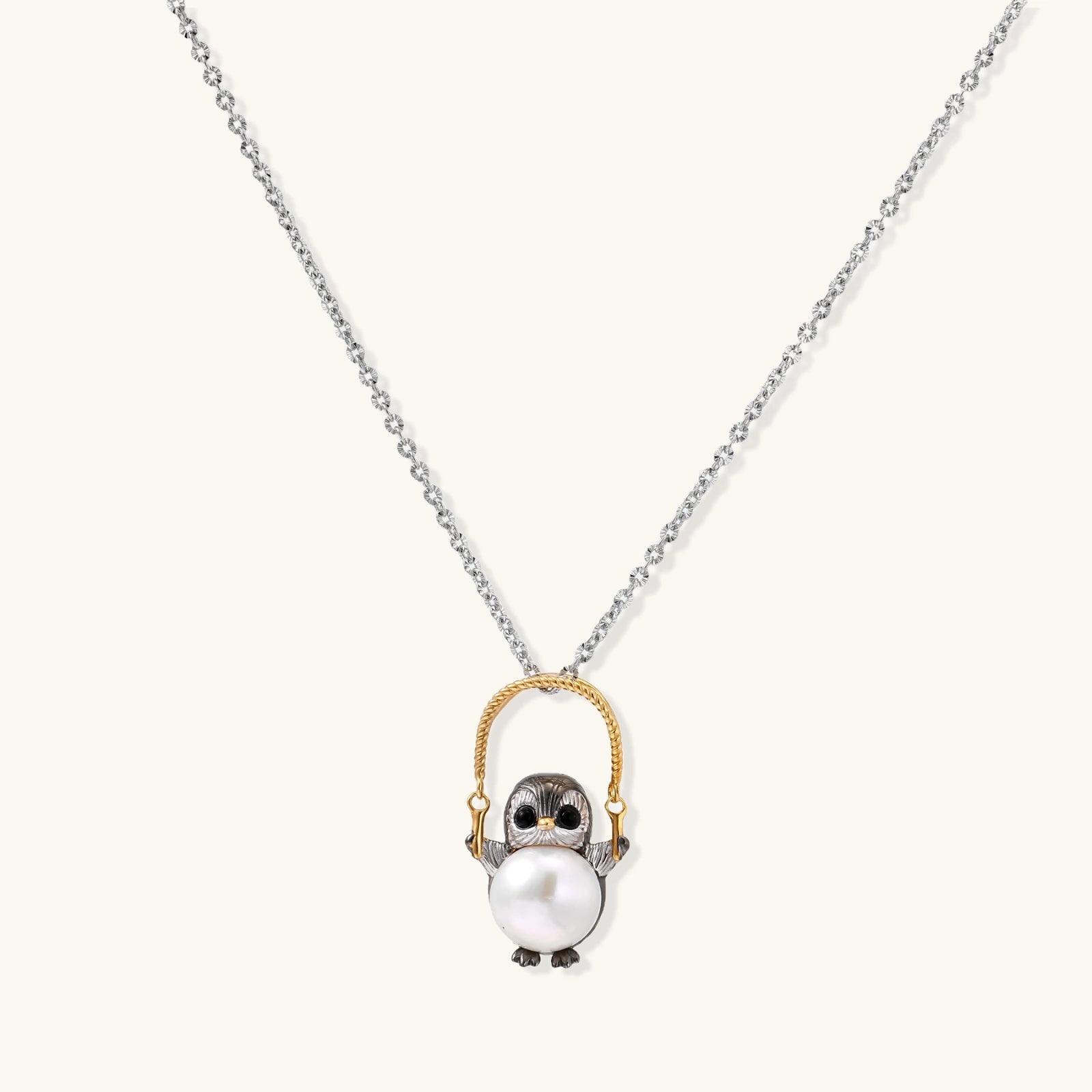 Whimsical penguin pendant holding a white pearl on a gold swing, rhodium-plated silver chain on cream background.