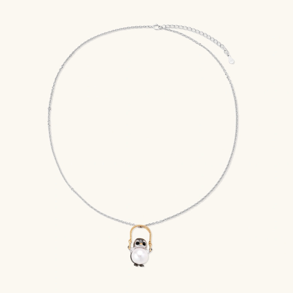 Whimsical penguin pendant holding a white pearl on a gold swing, rhodium-plated silver chain on cream background.