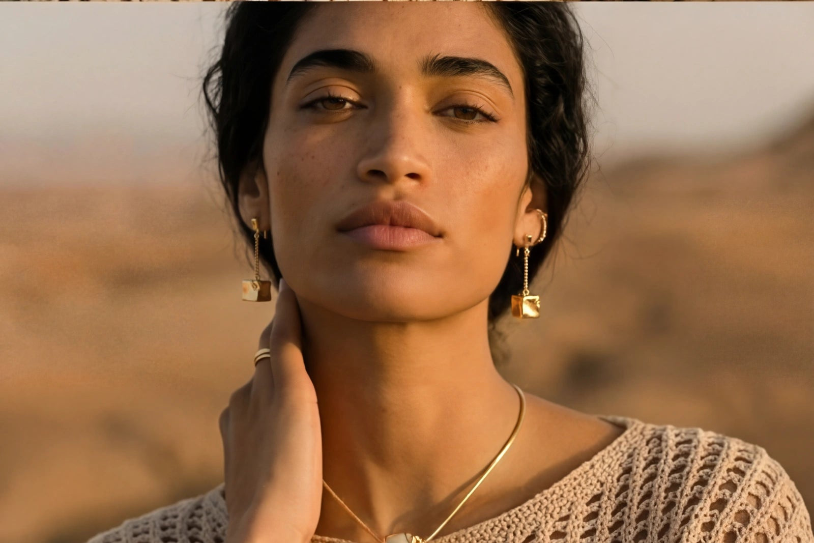 Woman wearing a beige knitted sweater and gold jewelry against a blurred natural background